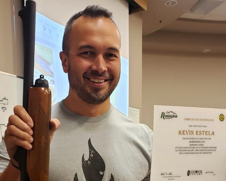 A man in a gray shirt with a shotgun and certificate standing in front of a screen