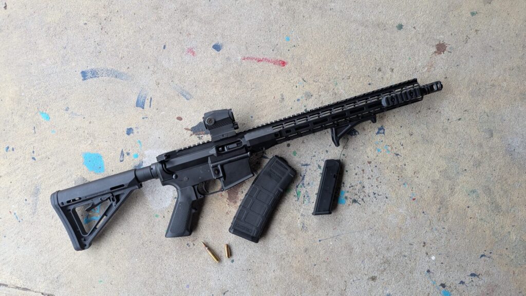 A black rifle with a red dot sight laying on concrete next to a rifle magazine, a handgun magazine, and two loose cartridges.