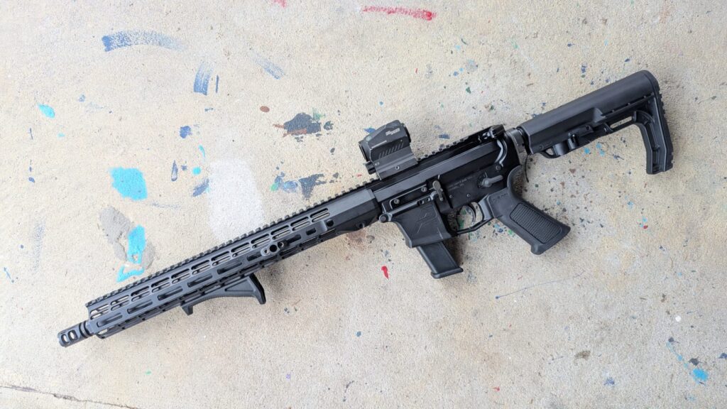 A full profile view of a black modern sporting rifle equipped with a red dot sight and a telescoping stock, resting on a speckled concrete surface.