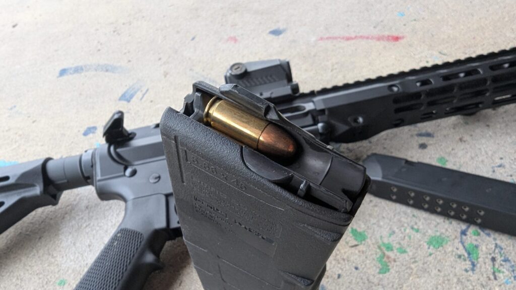A close-up of a loaded black rifle magazine in the foreground with a copper-jacketed round visible, and a rifle and handgun magazine blurred in the background.