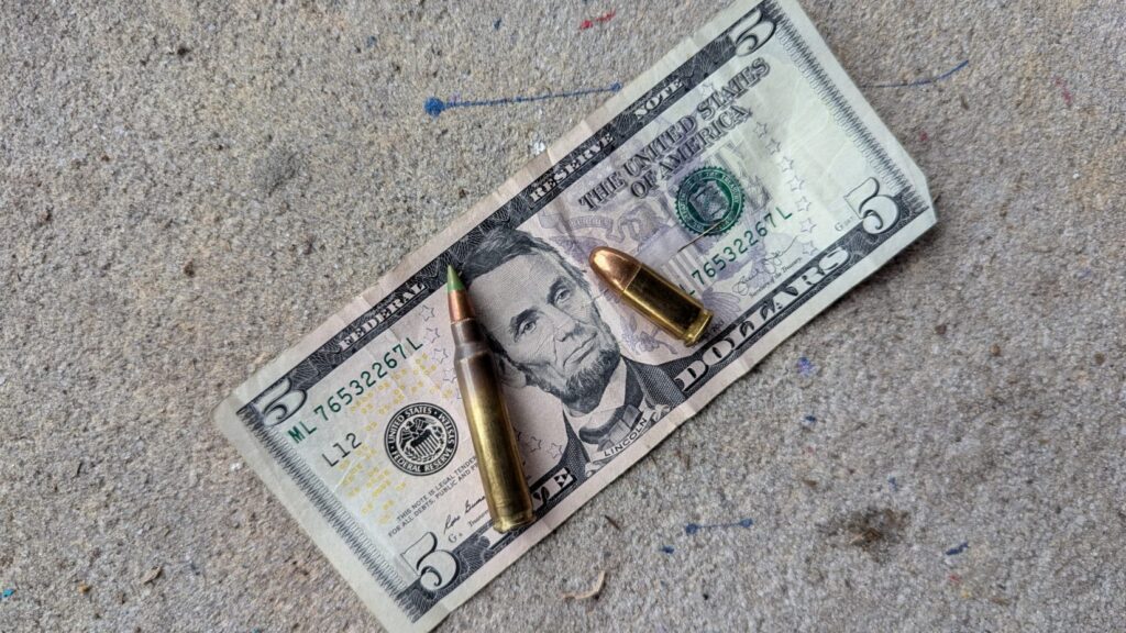 A five-dollar bill laying on the ground with a green-tipped rifle cartridge and a smaller copper-jacketed handgun cartridge resting on top of it.