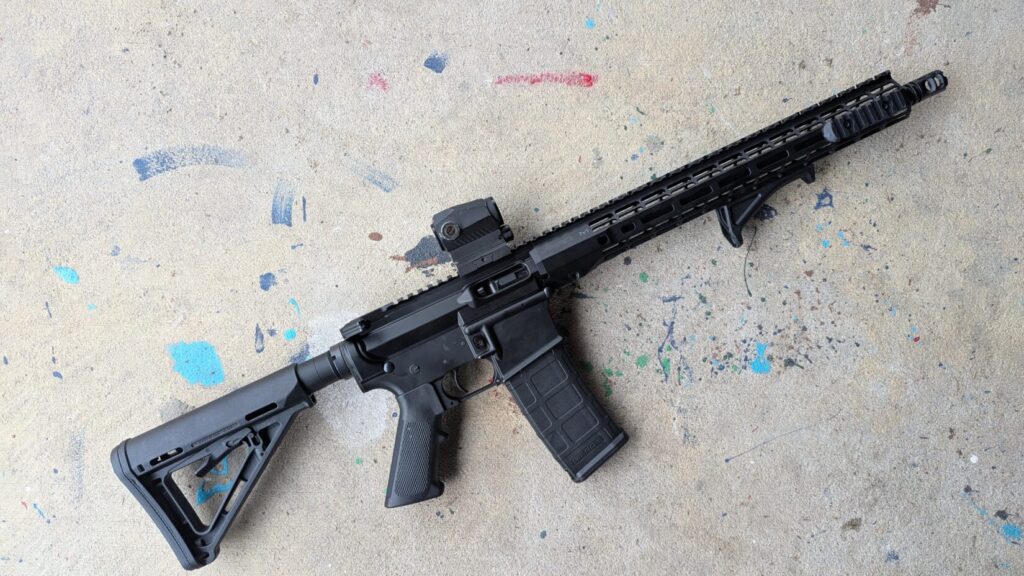 A black modern sporting rifle with a red dot sight and an angled foregrip, laying diagonally on a concrete floor with blue paint splatters.