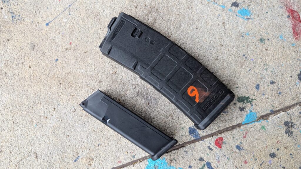 A black PMAG 30 rifle magazine marked with an orange "9" laying next to a smaller, slim black handgun magazine on a concrete surface.