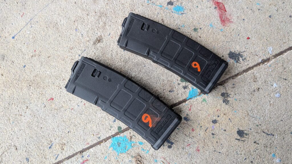 Two black PMAG 30 rifle magazines laying side-by-side on a concrete floor, each marked with a distinctive orange "9".