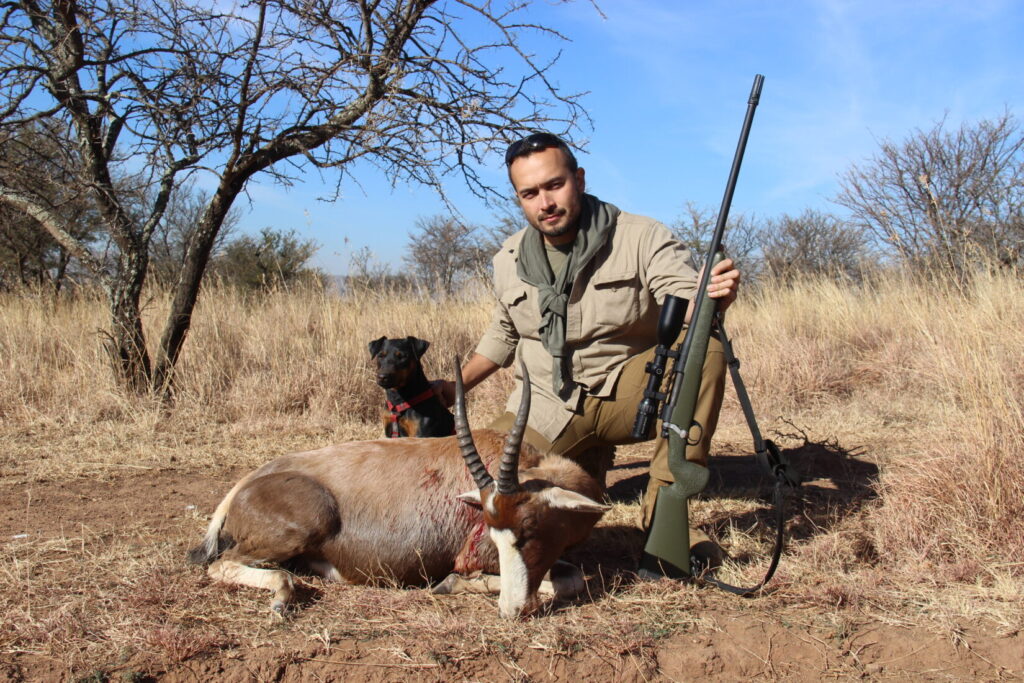 A man kneeling next to a blesbuck with a rifle and a hunting dog nearby in Africa.