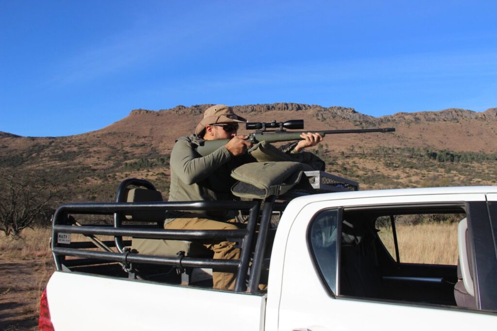 A man in the back of a safari truck taking aim with a rifle in the African landscape.