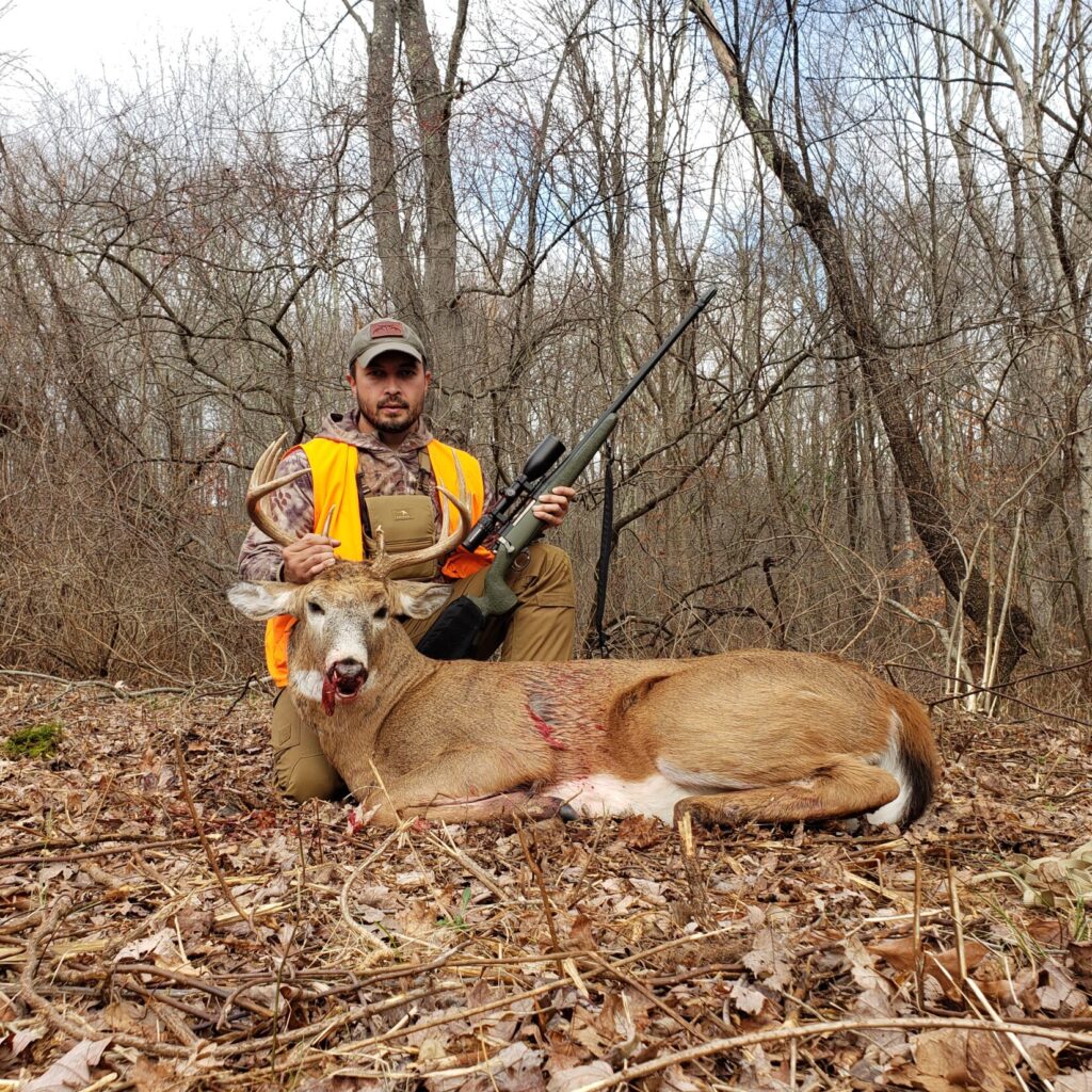 A man in a blaze orange vest next to a 10 point buck in the northeast woods
