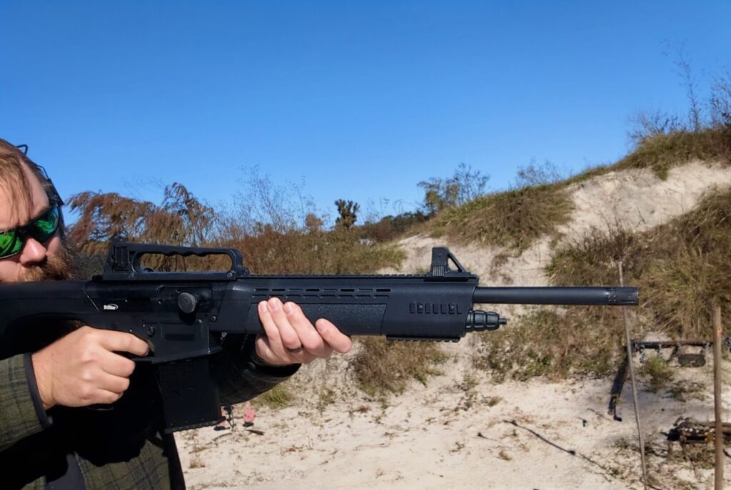 A man with a beard and green-tinted sunglasses aiming a black magazine-fed semi-automatic shotgun at an outdoor range.