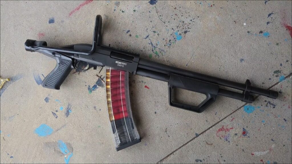 An SDS Imports Civet-12 pump-action shotgun with a folding stock and a transparent magazine loaded with red shells, resting on paint-splattered concrete.