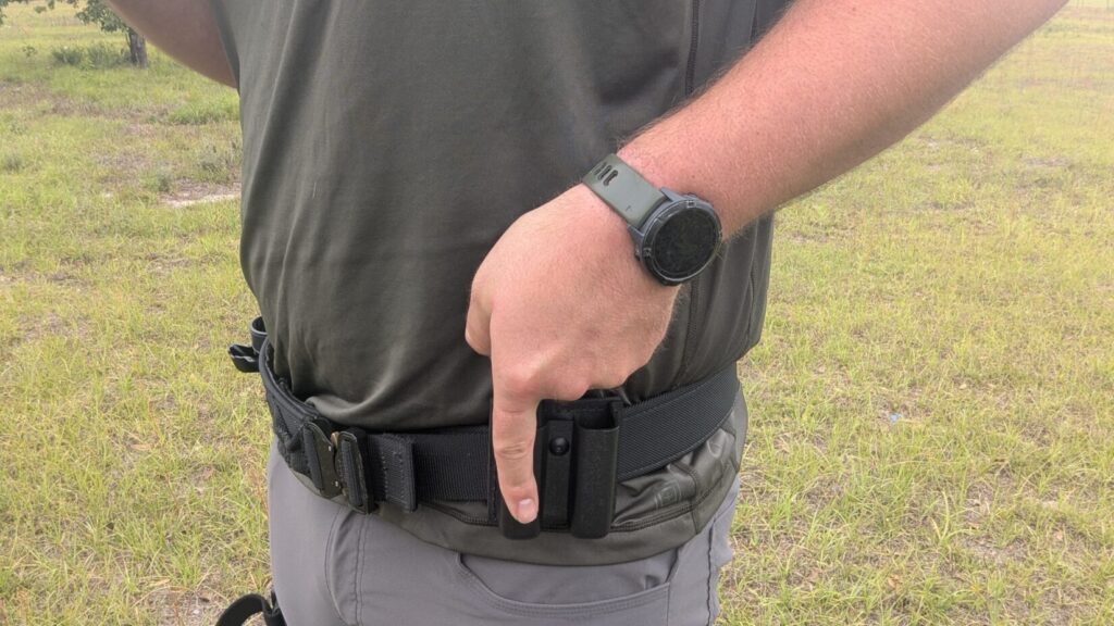 Close-up of a person's waist showing a black tactical belt with a spare magazine pouch and a smartwatch with an olive green band.