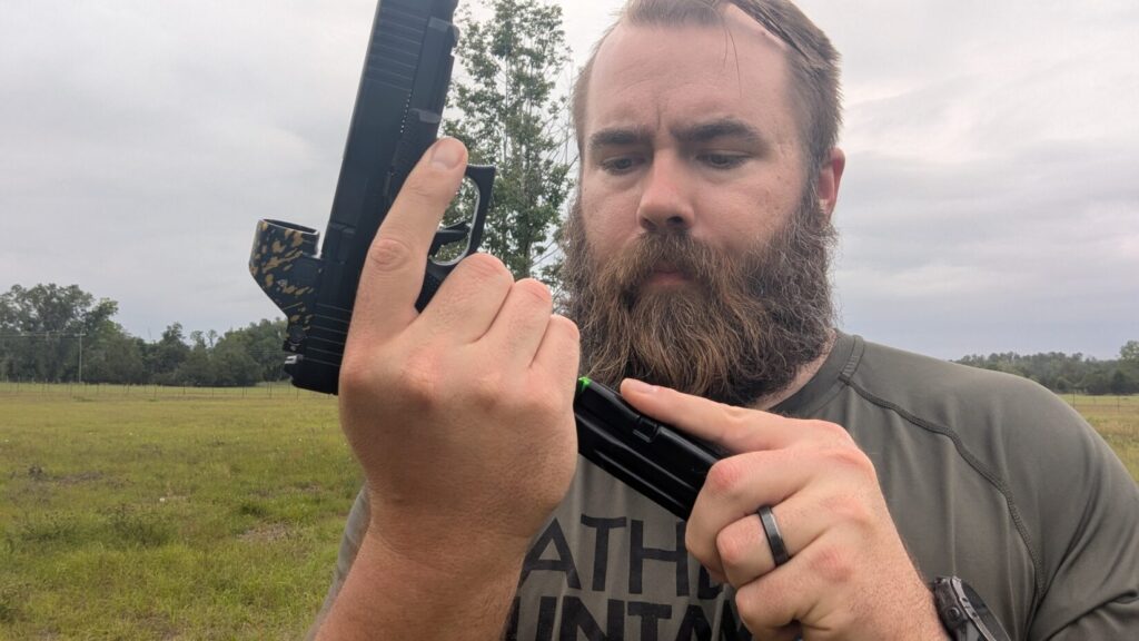 A man with a large beard wearing a grey shirt and a black ring, looking down while holding a black pistol and inserting a magazine.