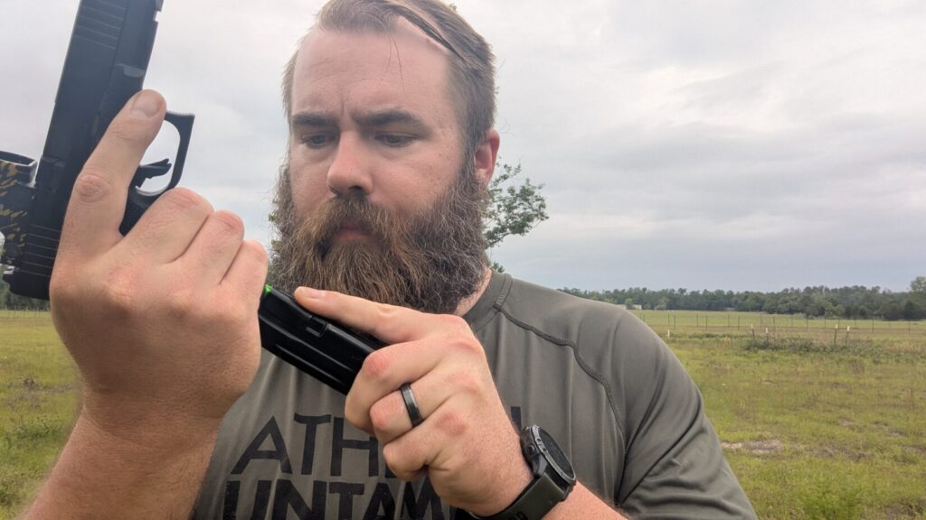 A man with a large beard wearing a grey shirt and a black ring, looking down while holding a black pistol and inserting a magazine.