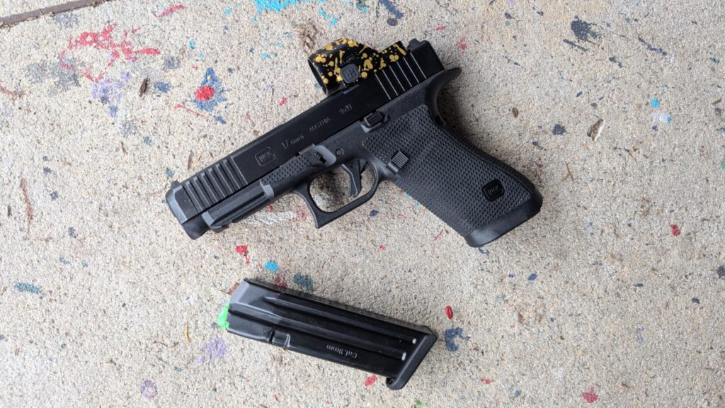 Top-down view of a black Glock 17 Gen 6 pistol with a gold-flecked red dot sight and a detached 9mm magazine on a speckled concrete surface.
