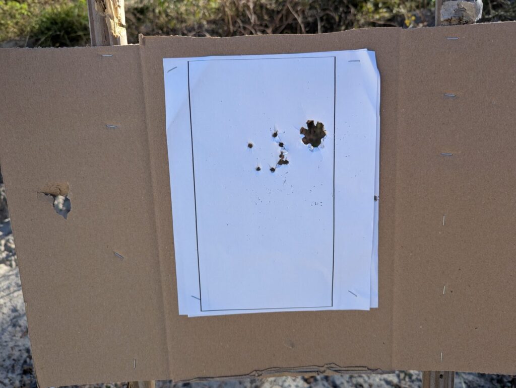 A white paper target stapled to a cardboard backing showing a tight grouping of several projectile impacts clustered in the upper center.