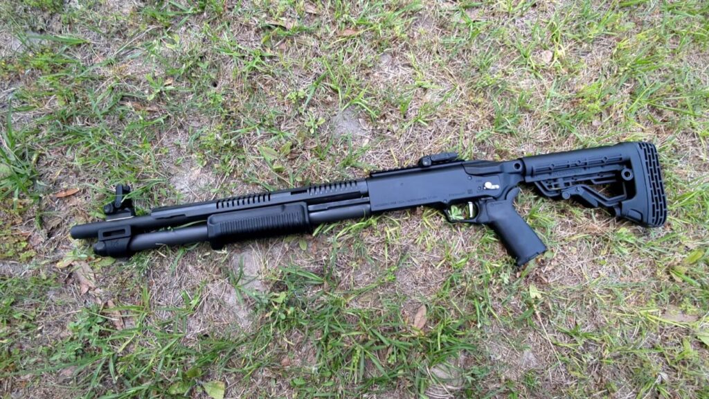 A black Mossberg 590 pump-action shotgun with an adjustable stock and pistol grip lies on a patch of grass.