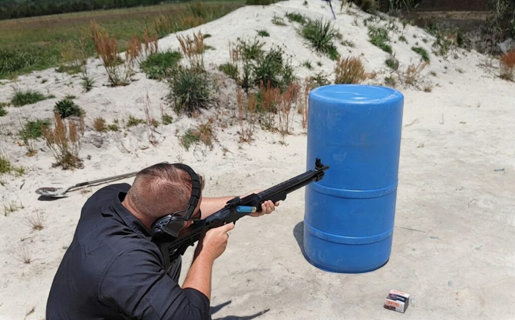 A person wearing hearing protection aiming a black shotgun over a blue plastic barrel at an outdoor shooting range.