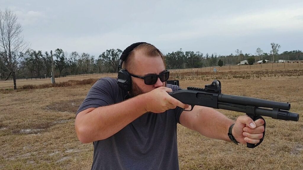 A close-up of a person in a gray shirt aiming a short-barreled shotgun with a red dot sight and a front vertical grip.