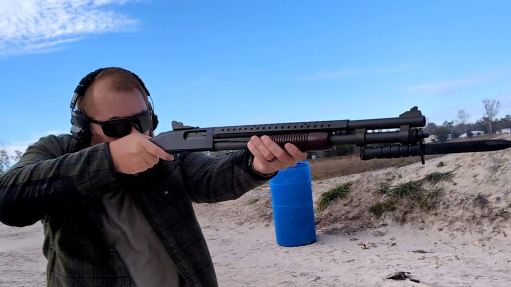 A bearded man in a plaid jacket aims a short-barreled pump-action shotgun with a wood forend at a firing range.