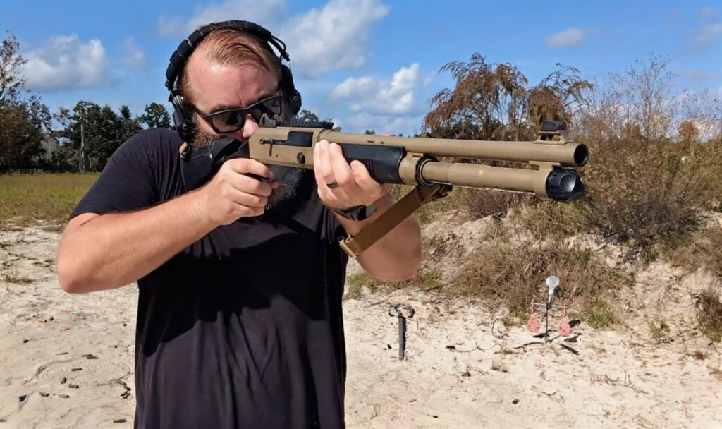A man wearing black headphones and sunglasses aims a tan-colored semi-automatic shotgun toward a target at an outdoor range.