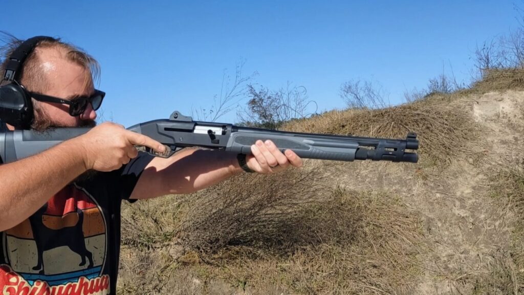 A man in a graphic t-shirt holds a grey Savage Renegauge shotgun, highlighting the receiver and extended magazine tube.