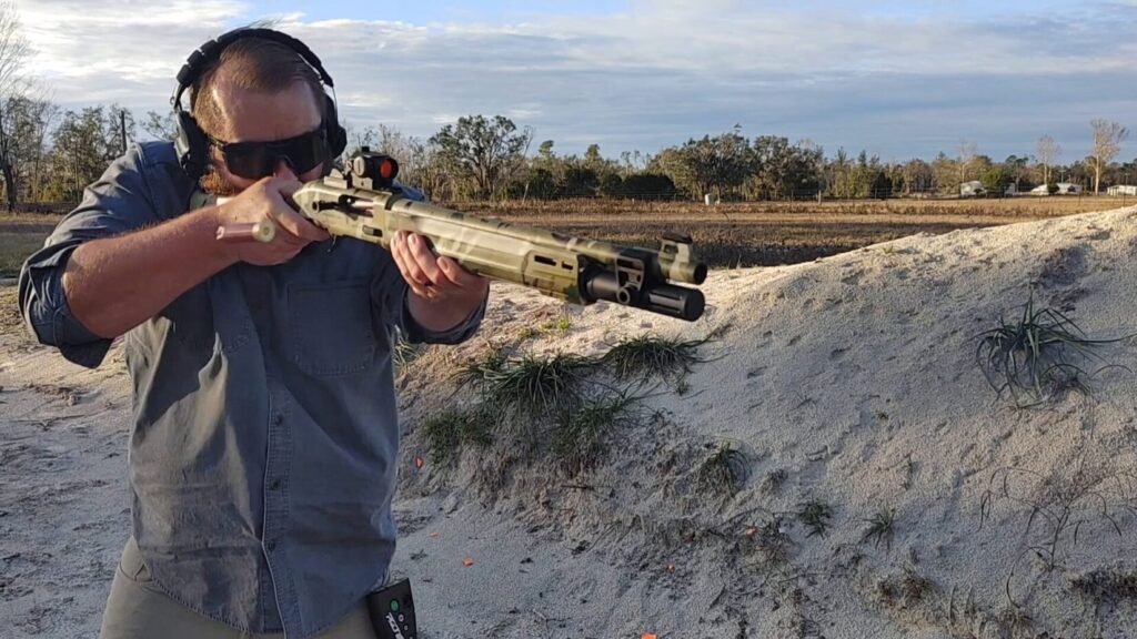 A person in a blue button-down shirt firing a camouflage-patterned semi-automatic shotgun, with a spent shell casing ejecting.