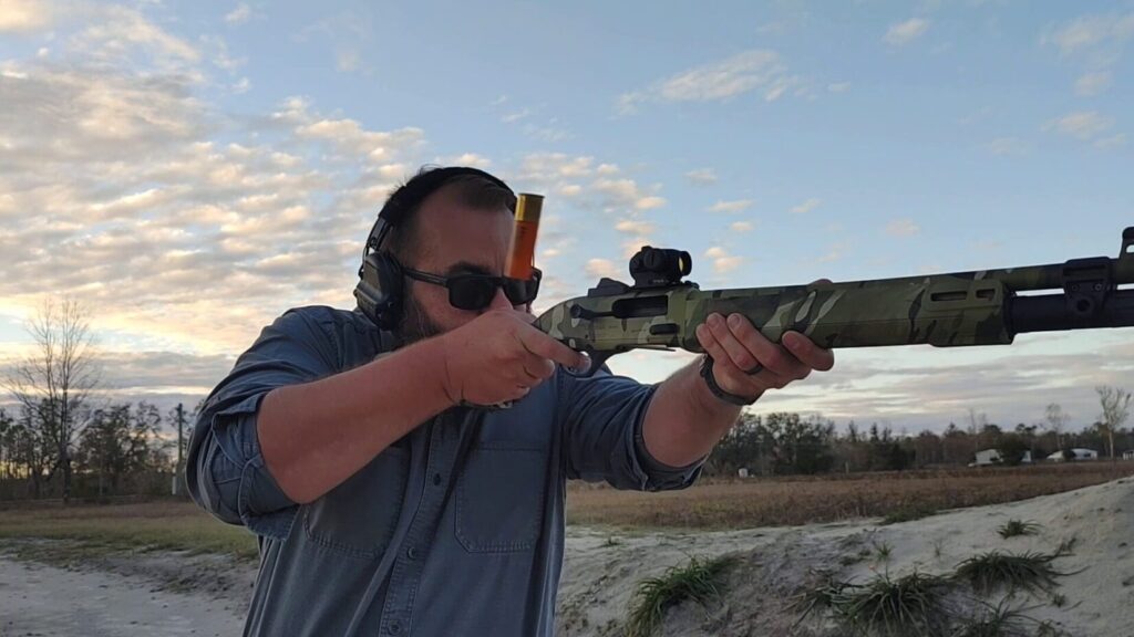 A person in a blue shirt aiming a camouflage semi-automatic shotgun directly toward the camera, with a shell in mid-air