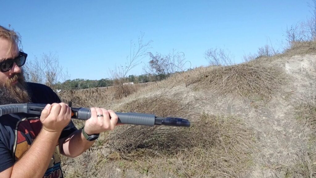 A person with a beard and sunglasses holding a modern shotgun with a gray forend and a prominent charging handle.