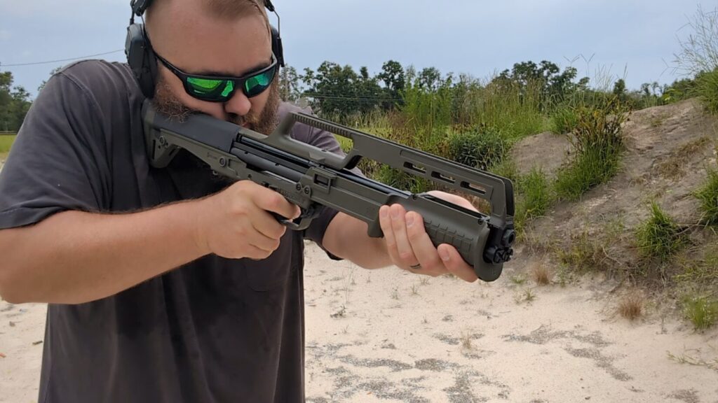 A man in a grey t-shirt and sunglasses aiming an OD green Kel-Tec KSG bullpup shotgun at an outdoor shooting area.
