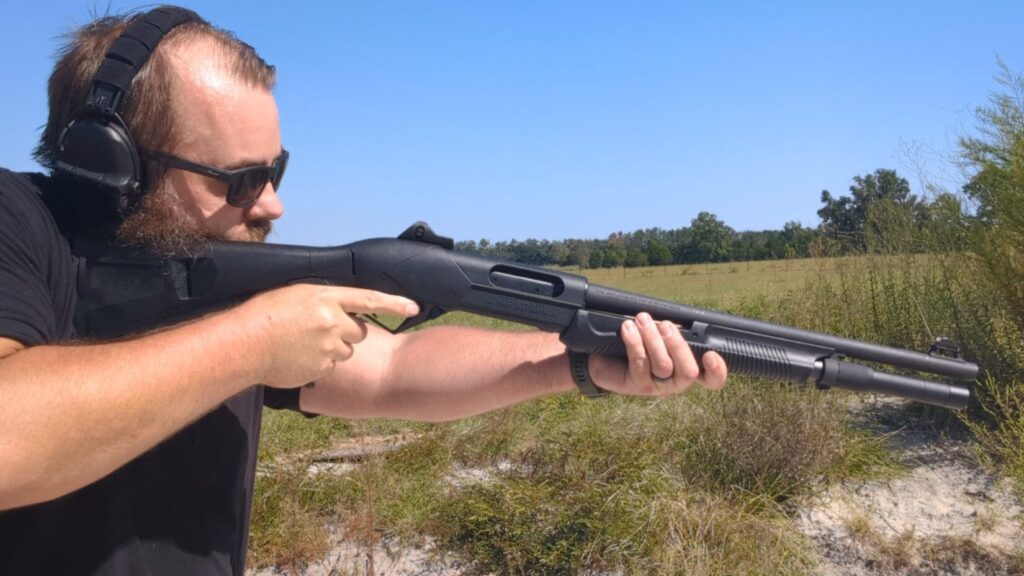 A man with a beard and sunglasses aims a black Benelli pump-action shotgun at an outdoor field.