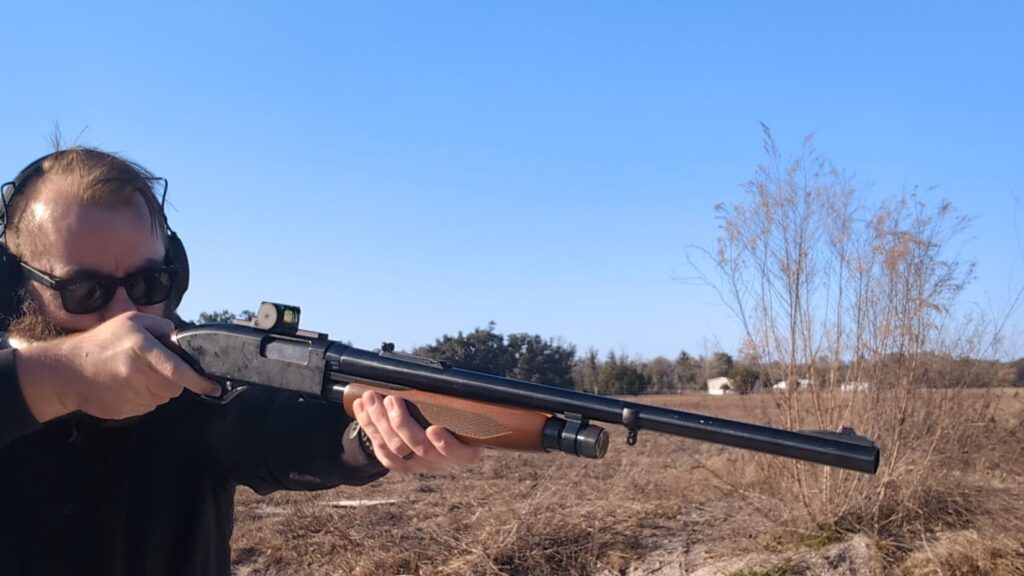 A man with a beard and sunglasses aims a black shotgun equipped with a red dot sight and wood forend at an outdoor range