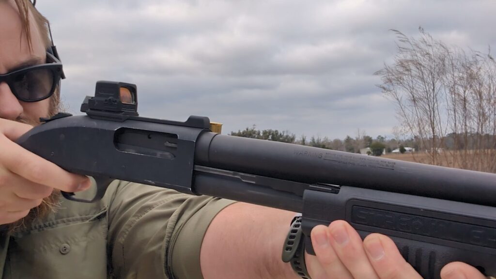 A close-up view focusing on the receiver and Holosun optic of a black Mossberg 590 shotgun, highlighting the integrated Streamlight TL-Racker forend light