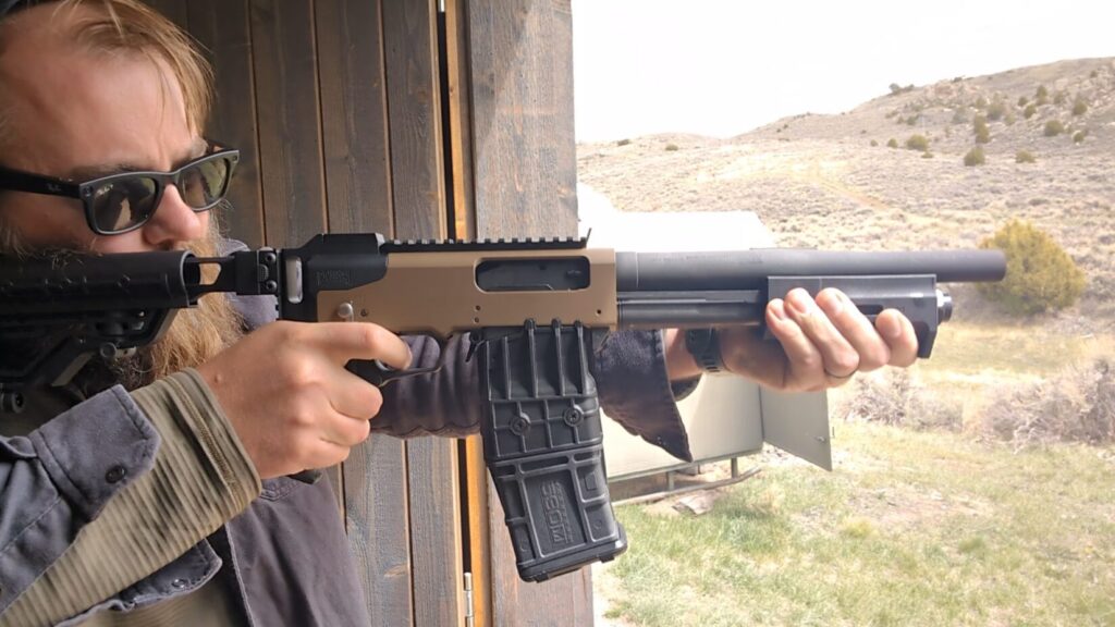  A close-up of a man aiming a tan and black Mossberg 590M magazine-fed shotgun equipped with an adjustable stock.