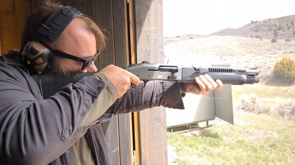 A man in a grey shirt aims a grey semi-automatic shotgun from within a wooden shooting stall.