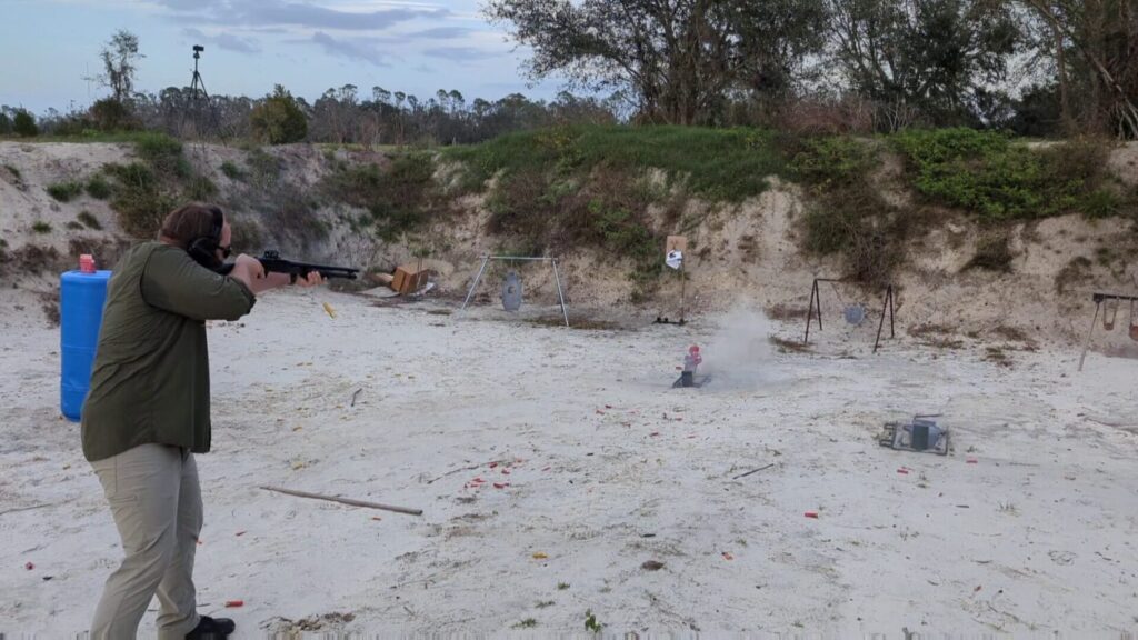 A wide shot of a person in an olive shirt firing a shotgun at a target range with steel plates and a sand berm.