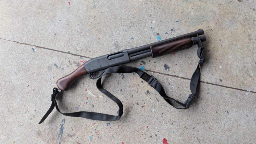 A Remington 870 Tac-14 with a wood raptor grip and a black carry sling, lying on a paint-flecked surface.