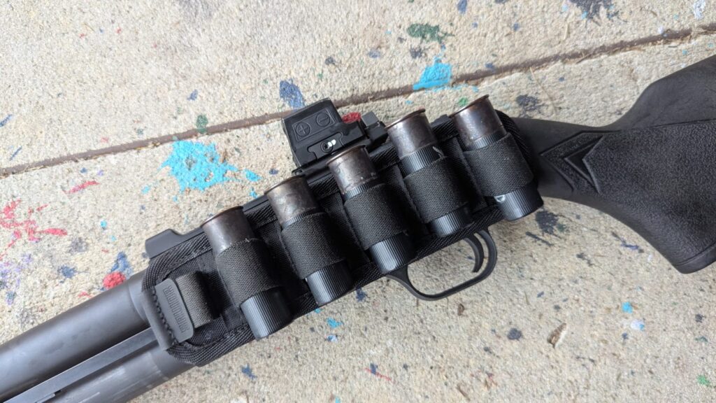 A close-up of a shotgun receiver featuring a side-mounted shell carrier holding five black shells, alongside a small red dot optic.