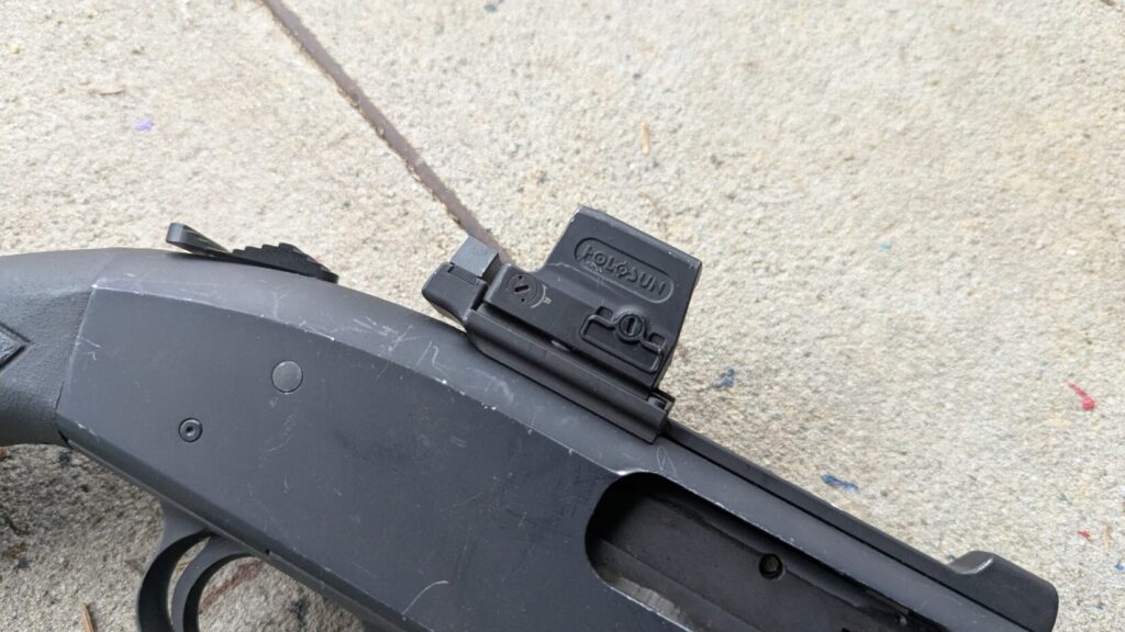 A high-angle close-up of a Holosun red dot sight mounted on the receiver of a black pump-action shotgun.