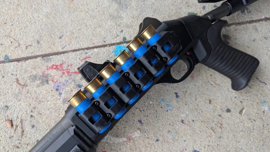 A close-up view of a black shotgun receiver with a side-saddle carrier holding six blue 12-gauge shells.