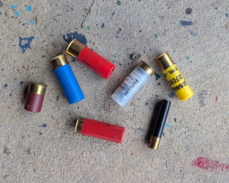 A variety of colorful shotgun shells, including blue, red, white, yellow, and black, scattered on a paint-splattered concrete surface.
