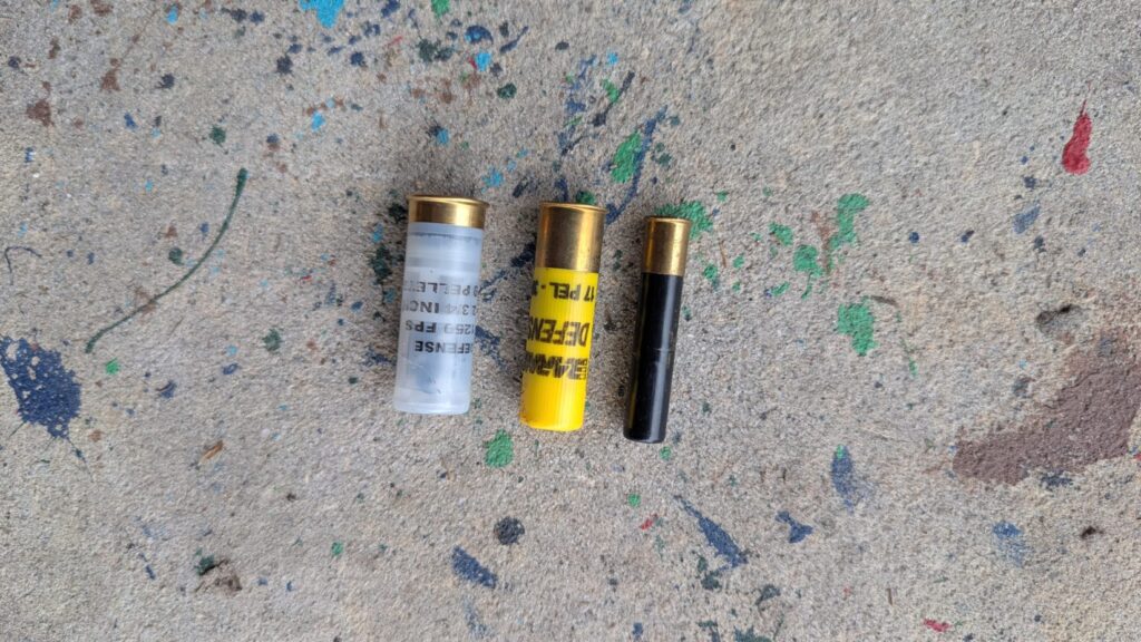 Three shotgun shells—one black, one yellow, and one clear—arranged vertically on a grey, paint-flecked concrete background.