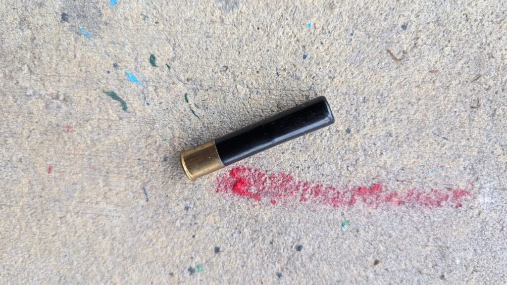 A single black shotgun shell with a brass base positioned on a grey concrete surface near a red paint streak.