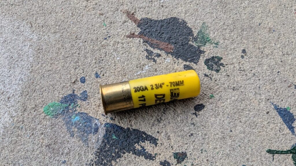 A single yellow 20-gauge shotgun shell with visible technical markings lying horizontally on paint-splattered concrete.
