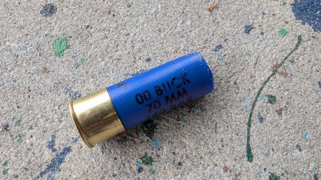 A close-up view of a single blue 12-gauge 00 buckshot shell lying on a textured, paint-splattered concrete surface.