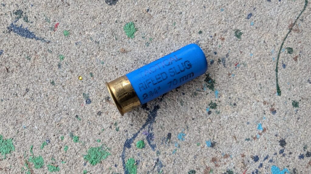 A close-up view of a single blue 12-gauge 00 buckshot shell lying on a textured, paint-splattered concrete surface.