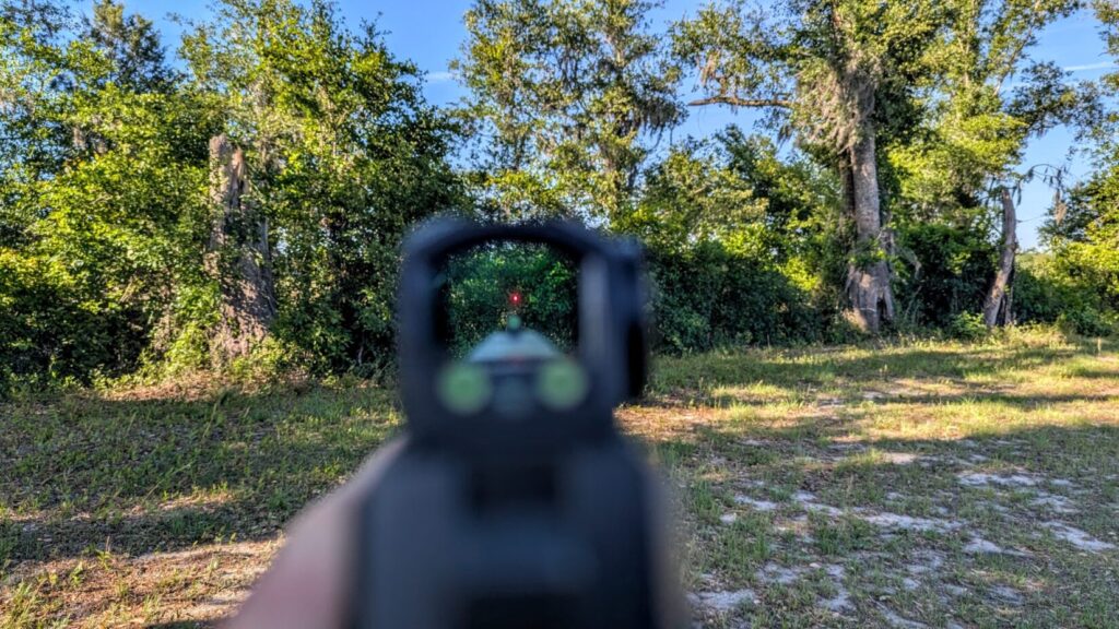 A "sight picture" view looking through the Steiner MPS-C window, showing the red dot reticle aligned with iron sights against a background of trees.