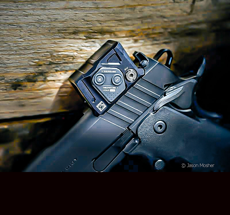 A close up of the COA red dot mounted on a black gun, leaning against a wood wall.