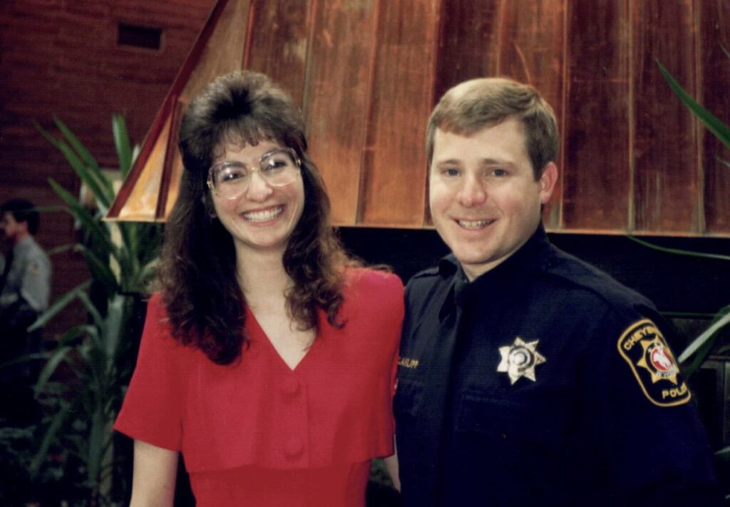 Photo of a lovely woman in a red dress with large glasses and big 1990s hair, with a recent police academy graduate.