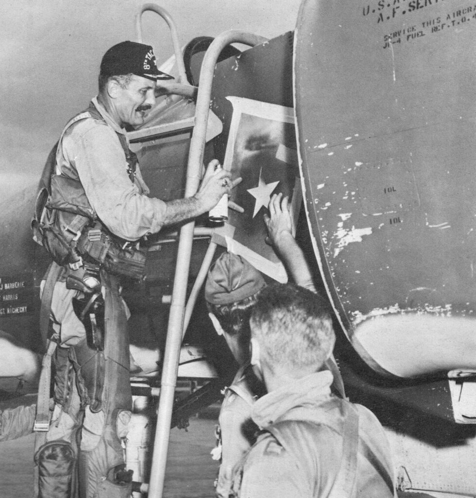 Black and white photo of Colonel Robin Olds spray painting over a star-shaped stencil on the side of an F-4 Phantom II, signifying his arial victory over a MiG on May 4, 1967.