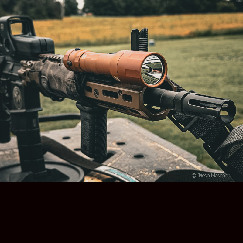 A brown cloud defensive REIN 3.0 weapon-mounted lighton a rifle, sitting on a table in a field.