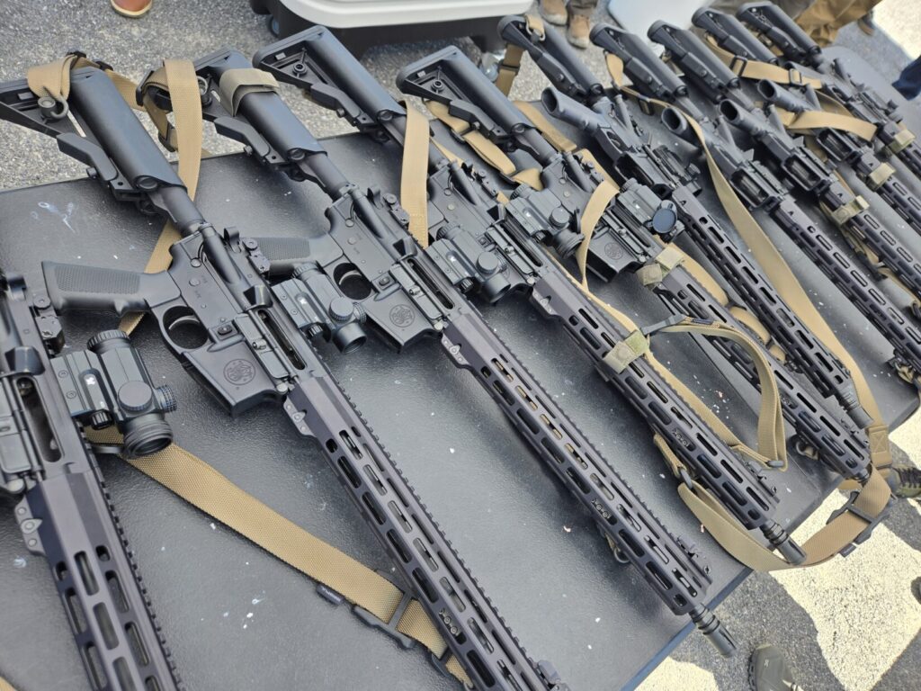 Multiple AR 15 rifles with red dot optics and slings on a table